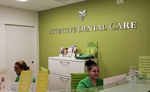 An interior view of a dental clinic reception area with a sign reading Attentive Dental Care and two individuals present, one seated at a desk and another standing.